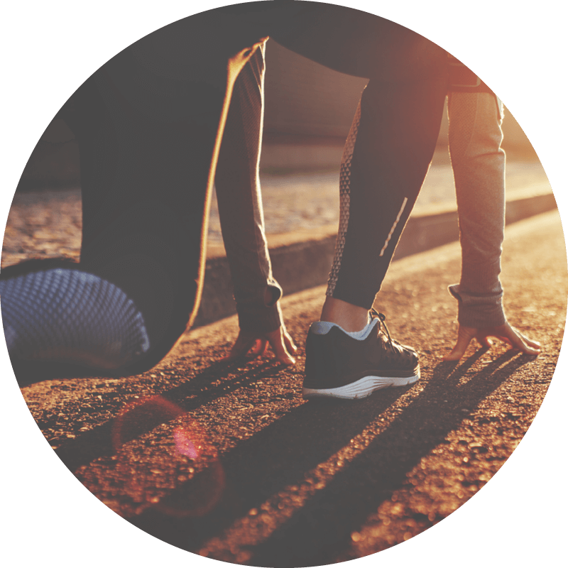 Person crouching in a starting position, wearing athletic shoes, on a sunlit road surface, preparing to run.